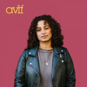 A person with curly hair, wearing a black leather jacket and gray shirt, stands against a pink background. They have a calm expression and are wearing hoop earrings and a necklace. The letters 