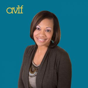 A woman with shoulder-length hair smiles against a teal background, wearing a dark sweater over a grey top with a necklace. The letters 