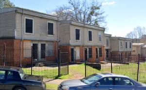 A row of two-story townhouses with boarded-up windows and doors, surrounded by a black metal fence. Two parked cars are visible in front, and leafless trees can be seen in the background under a clear blue sky.