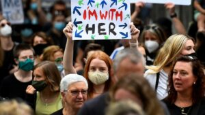 A crowd of people, many wearing masks, gathers at a protest. A red-haired person in the center holds up a sign reading 