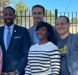 Four people standing in front of a metal fence outdoors. One is wearing a suit and blue tie, another is in a casual gray blazer, one is in a light gray jacket, and the fourth person is wearing a white and black striped sweater. Trees and a clear sky are in the background.