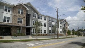 Street view of a modern, three-story apartment building with a combination of brick and gray siding. The road in the foreground is lined with trees, and there are power lines overhead under a partly cloudy sky.
