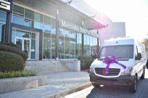 A white van with a large purple bow on the hood is parked in front of a Mercedes-Benz dealership. Sunlight is shining, casting a bright halo effect. The building is modern with glass windows and a sign saying 