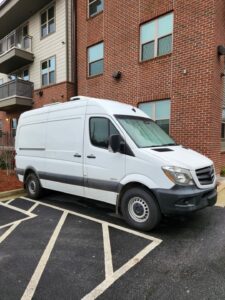A white cargo van is parked in a designated parking space next to a brick building with multiple windows. The building has balconies, and the surrounding area includes a sidewalk and some landscaping.