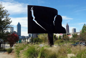Silhouette sculpture of a figure waving, set in a grassy area with a backdrop of an urban skyline on a clear day. Tall buildings and trees are visible, and the sky is blue with scattered clouds.