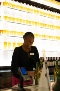 A person smiling behind a counter with wine bottles, against a wall of illuminated bottles. They are wearing a black jacket and holding a wine bottle with a shelf of more bottles in the background.
