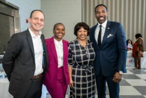 Four people stand together smiling in a well-lit room. Two men are on either side, wearing suits—one in a dark suit and one in light. Two women in the middle, one in a pink suit and the other in a checkered dress. A table with items is in the background.