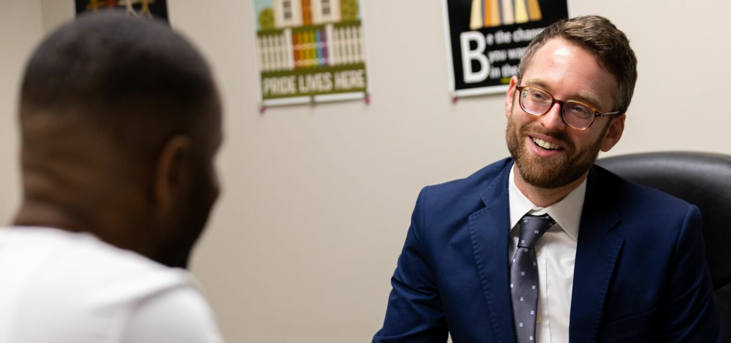 Two people are sitting at a desk in an office. One man in a blue suit and glasses is smiling, engaged in conversation. The other person, seen from behind, is wearing a white shirt. Various posters are on the wall in the background.