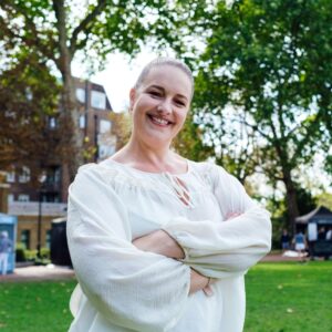A person with long hair tied back smiles confidently, standing with arms crossed in a park. They wear a light, flowy top. Trees and buildings are visible in the background on a sunny day.
