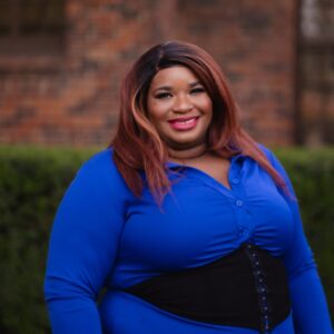 A woman with long, straight, reddish-brown hair, wearing a blue button-up shirt and a black waist belt, is smiling outdoors. A brick wall and green bushes can be seen in the background.