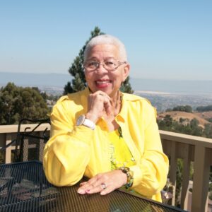 A woman with short gray hair and glasses, wearing a bright yellow jacket, smiles while seated at an outdoor table. Behind her is a scenic view of hills, trees, and a cityscape under a clear blue sky.
