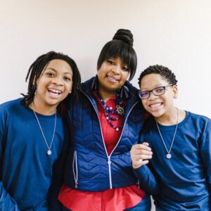 A smiling woman stands between two boys. She's wearing a blue quilted jacket over a red blouse with a beaded necklace. Both boys wear matching blue shirts and silver pendants. They are all posing in front of a plain, light-colored wall.