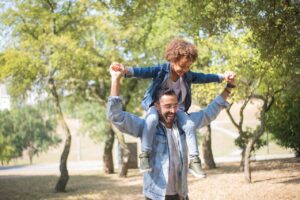 A man joyfully carries a child on his shoulders while walking in a sunlit park. The child is smiling, wearing a denim jacket and sneakers. Trees with green leaves surround them, creating a cheerful and peaceful atmosphere.