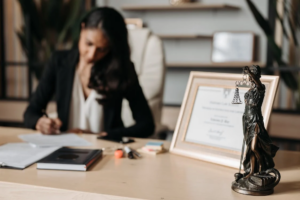 A woman sits at a desk, writing on paper. In the foreground, a statuette of Lady Justice is visible next to a framed certificate. The room has bookshelves and a plant in the background, suggesting a professional office setting.