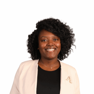 A woman with curly hair smiles at the camera, wearing a light-colored blazer over a black top and a necklace with a cross pendant, standing against a plain white background.
