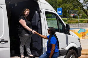 A woman in a blue shirt helps another woman step out of a white van. They smile at each other in a sunny outdoor setting with trees, a basketball court, and an accessibility parking sign in the background.