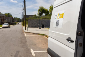 A white van is parked on a residential street next to a sidewalk with a stone wall. The van has a sign with the letters 