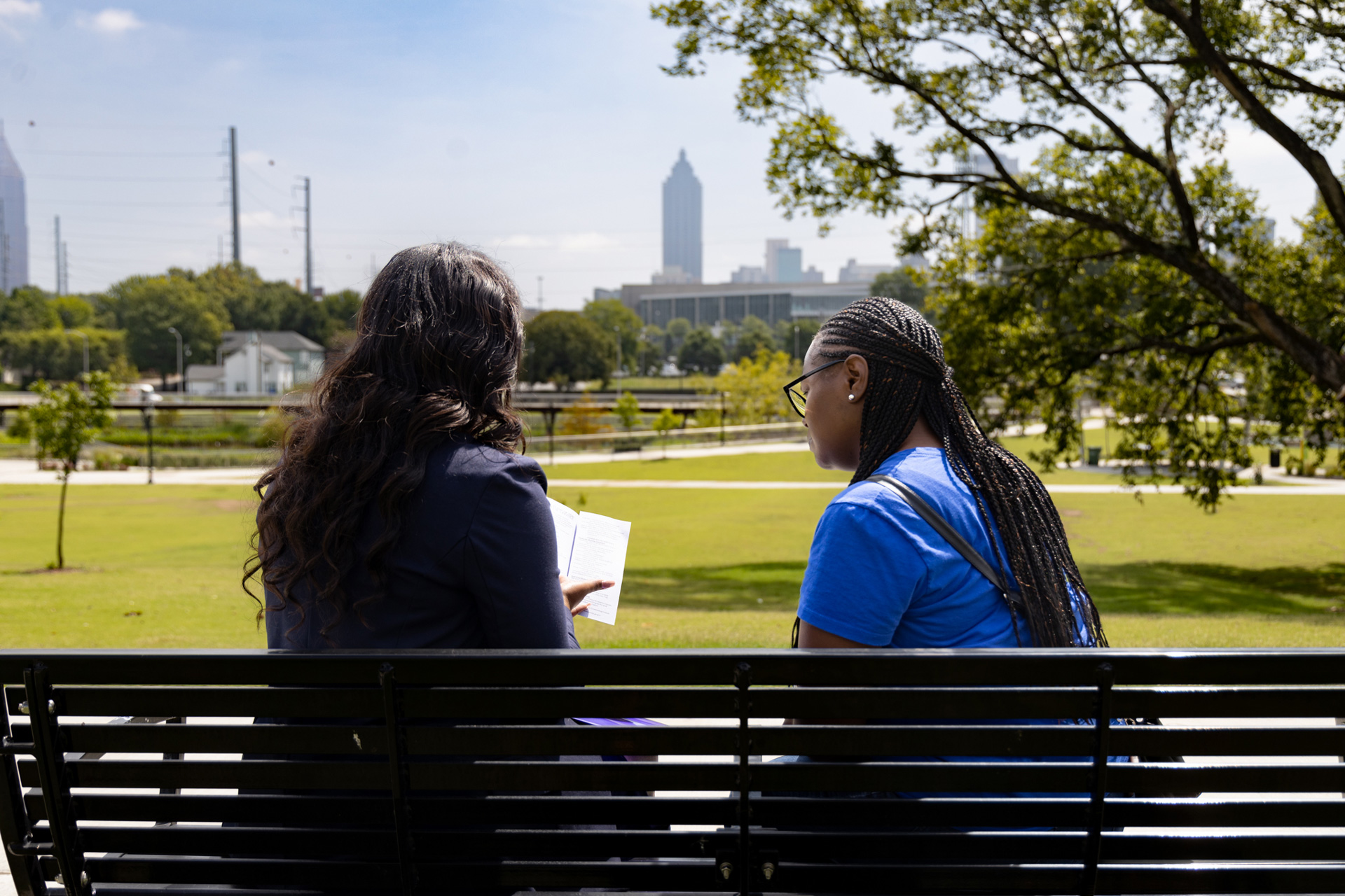 Two people sitting on a bench in a park, with one reading a book. They face away from the camera, looking towards a city skyline in the background. Trees and grass surround them on a sunny day.