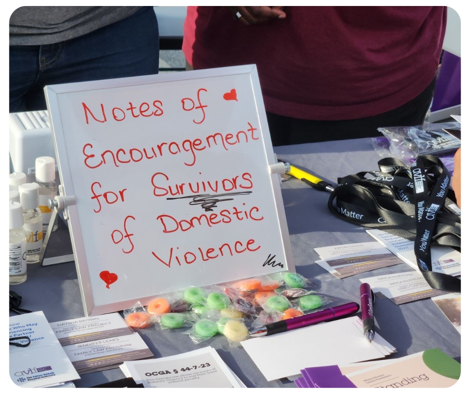 A table displays a sign reading "Notes of Encouragement for Survivors of Domestic Violence" surrounded by pamphlets, candies, pens, and items with motivational messages.