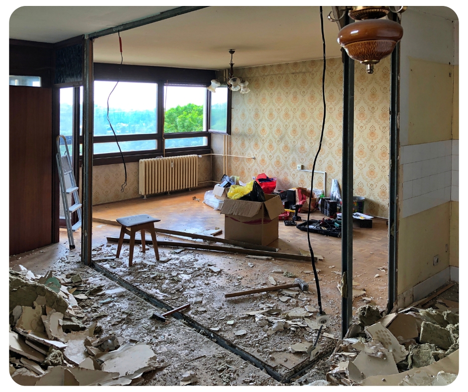 Room under renovation with a partially demolished wall in the foreground. Debris is scattered on the floor. The space is cluttered with boxes and tools. Large windows reveal trees and a distant view outside. Vintage wallpaper covers one wall.