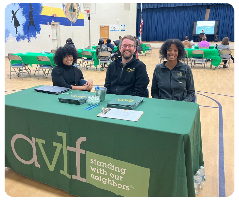 Three people sit at a table covered with a green cloth displaying "avlf standing with our neighbors." They are indoors at a community event, with tables and chairs in the background.