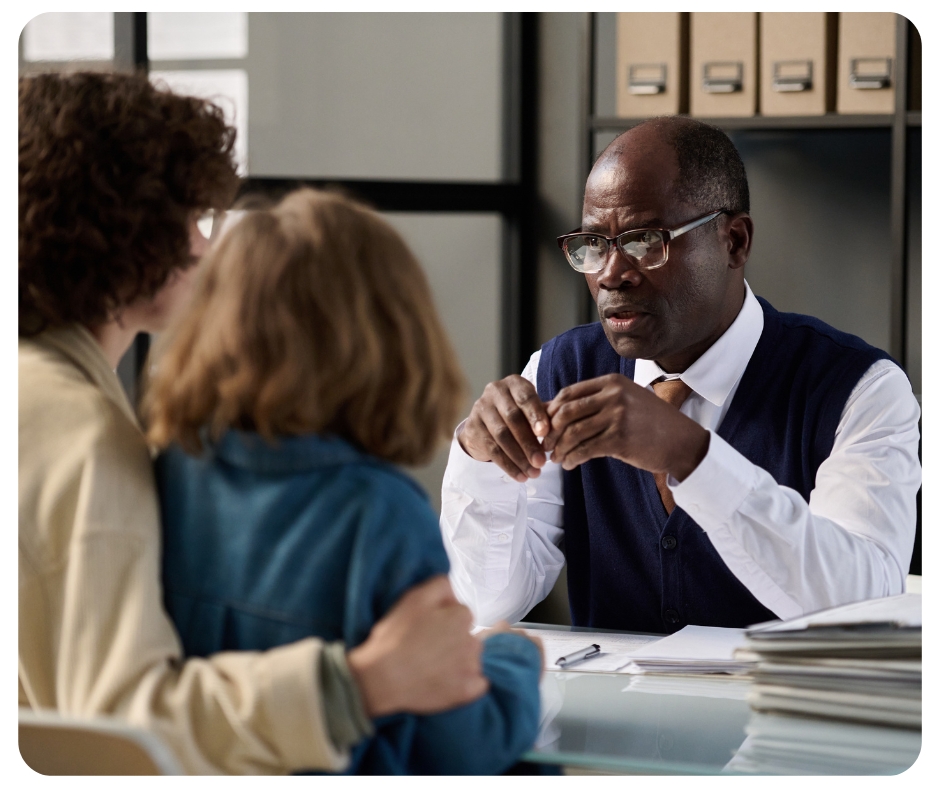 A man in glasses and a blue vest sits at a desk talking to two people. They appear to be in an office setting. The desk is covered with papers and a stack of folders. The two people are seen from behind, suggesting a professional consultation.