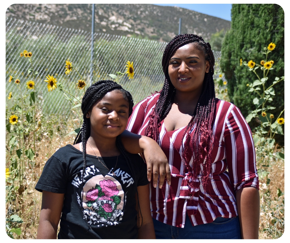 A woman and a young girl stand together in a field of sunflowers. The woman is wearing a striped shirt, and the girl has a black T-shirt. A chain-link fence and a grassy hill are in the background.