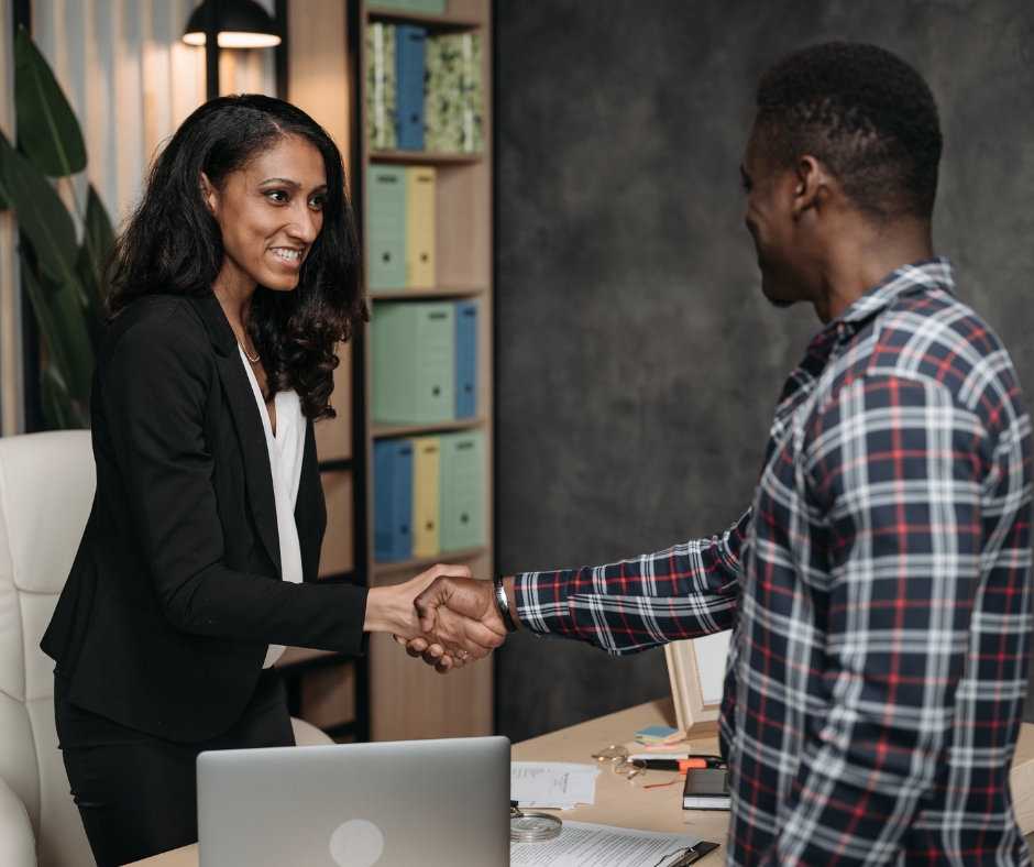 A woman in a black suit and a man in a plaid shirt are shaking hands across a desk in an office setting. The desk has a laptop and documents, and a shelf with colorful binders is in the background.