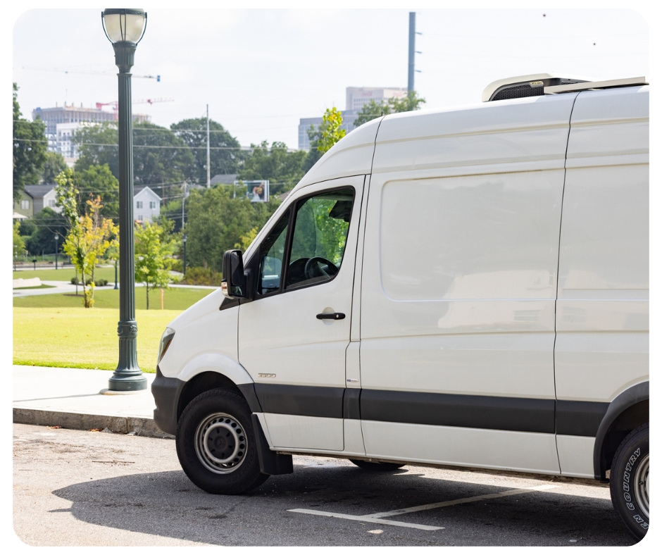 A white delivery van is parked on a street near a grassy park with trees. A lamppost stands beside the van, and buildings are visible in the background under a clear sky.