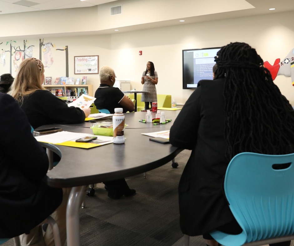 A group of people seated at a table in a classroom setting, listening to a speaker standing near a screen. The room has brightly colored chairs and a painted wall in the background. Papers and bottles are on the table.