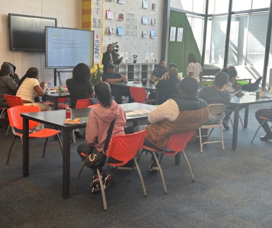 A group of people sit at tables in a classroom, facing a presentation screen. The presenter stands near the screen, gesturing. The room has large windows and a bright atmosphere.