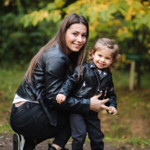 A woman and young child, both wearing black leather jackets, smile outdoors in a park. The woman is crouching and holding the child, who looks excited. They are surrounded by green foliage and trees.