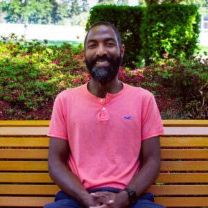 A man with a beard and a bright smile sits on a wooden bench in a garden. He wears a pink shirt and blue pants. Behind him, green bushes and pink flowers are visible, creating a colorful and serene backdrop.