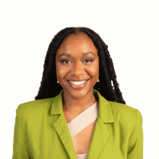 A woman with long, curly black hair smiles at the camera. She is wearing a bright green blazer over a tan top and has a friendly expression, posed against a plain white background.