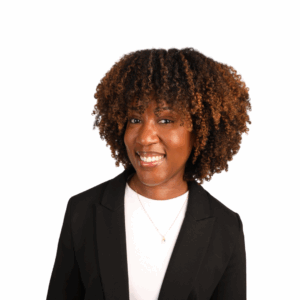 A woman with curly, shoulder-length hair and a warm smile wears a black blazer over a white top, standing against a plain white background.