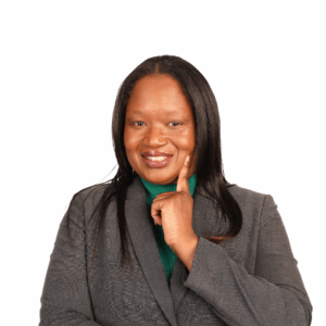 A woman with long dark hair wearing a gray blazer and green top, smiling and touching her chin with one finger, posing confidently against a plain white background.