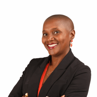A smiling woman with a shaved head, wearing a black blazer over a red top, gold earrings, and a necklace, standing with arms crossed against a white background.
