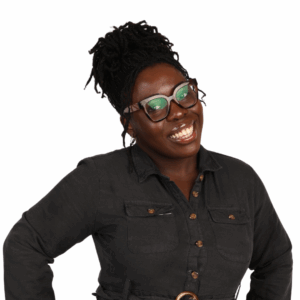 A smiling woman with dark skin and braided hair in a bun, wearing glasses and a dark button-up dress, stands against a white background with one hand on her hip.