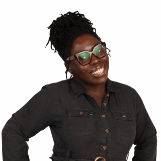 A smiling woman with dark skin and braided hair in a bun, wearing glasses and a dark button-up dress, stands against a white background with one hand on her hip.