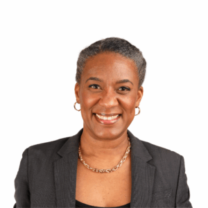 A middle-aged woman with short, gray hair smiles at the camera. She is wearing a dark blazer, a black top, gold hoop earrings, and a gold chain necklace, posed against a plain white background.