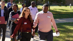 A group of professionally dressed individuals walking outdoors on a sunny day. A woman in a red blouse and a man in a pink sweater, both with name tags, lead the group, carrying folders and papers. Green grass and trees line the background.