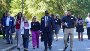 A group of people walking outdoors on a sunny day. Some are dressed in business attire, and others in casual outfits. They are carrying bags and folders, and trees are visible in the background.