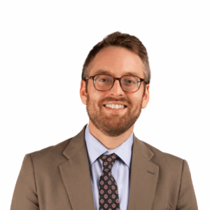 A smiling man with short brown hair, a beard, and glasses wearing a tan suit, light blue shirt, and patterned tie, standing against a plain white background.