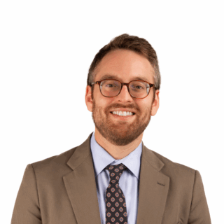 A smiling man with short brown hair, a beard, and glasses wearing a tan suit, light blue shirt, and patterned tie, standing against a plain white background.