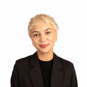 A person with short blonde hair, wearing small hoop earrings, a black shirt, and a black blazer, smiles slightly while facing the camera against a plain white background.