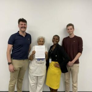 Four people stand against a white wall. An older woman in a white shirt holds a document. The man on the left wears a navy shirt, and the two people on the right wear a black jacket with a yellow skirt and a maroon sweater, respectively. They all smile.