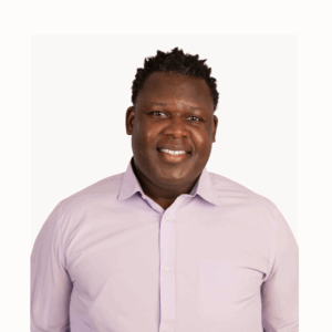 A man with short curly hair smiles at the camera. He is wearing a light purple button-up shirt and is posed against a plain white background.