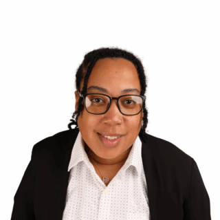 A person with medium skin tone, wearing glasses, a white polka-dotted shirt, a black blazer, and a necklace, smiles at the camera against a plain white background.