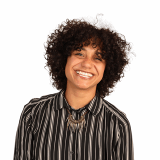 A person with curly dark hair smiles brightly, wearing a black and white striped shirt and a statement necklace, standing against a plain white background.