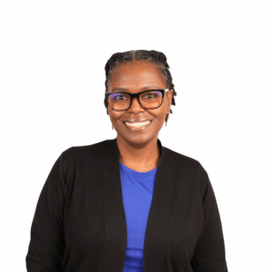 A smiling woman wearing black glasses, a blue shirt, and a black cardigan stands in front of a white background.